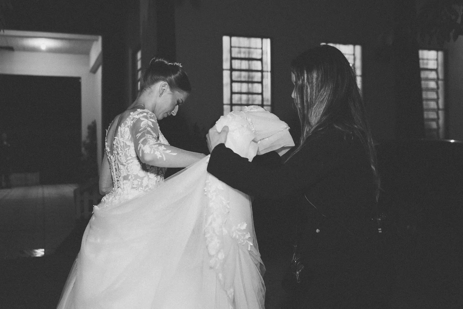 Camila ajudando a noiva a segurar o vestido, em preto e branco